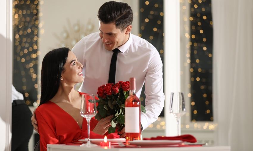 Junges Liebespaar am Esstisch - der Mann übergibt seiner Partnerin rote Rosen - Schöne Ideen zum Jahrestag