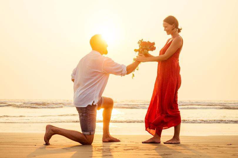 Paar am Strand - Mann auf Knien überreicht Rosenstrauß - Ideen zur Rosenhochzeit